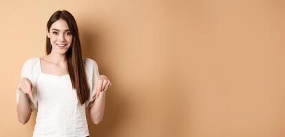 Cheerful young girl with lovely smile, pointing fingers down, making announcement or inviting click link, showing logo, beige background photo