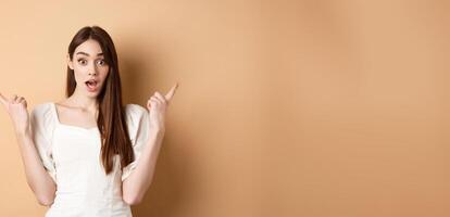 Surprised young girl pointing sideways at left and right, gasping and saying wow amazed, showing two cool choices, beige background photo