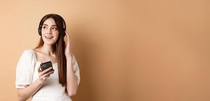 Attractive girl listening music in wireless headphones, holding mobile phone and looking aside with dreamy smile, beige background photo