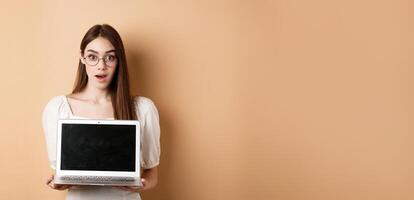 Surprised young woman in glasses showing laptop screen with amazed face, standing on beige background photo