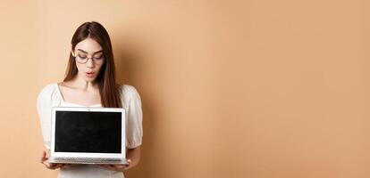 Excited girl in glasses show laptop screen, demonstrate online offer on computer, standing against beige background photo
