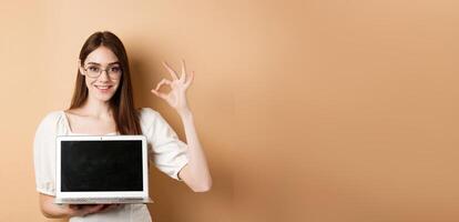E-commerce. Smiling young woman in glasses showing okay sign and laptop screen, recommending internet promo, standing on beige background photo