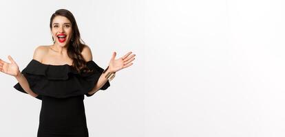 Beauty and fashion concept. Image of surprised and happy young woman in party dress reacting to good news, raising hands up and smiling amazed, white background photo