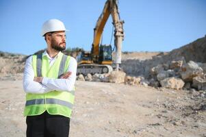 Extraction of stone. Male worker next to stone quarry. Engineer at construction site. Stone quarry worker with papers. He's calling someone on phone, excavator with bucket in background. photo