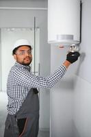 Indian plumber using screwdriver while fixing boiler or water heater, working on heating system in apartment photo
