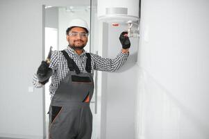 Indian plumber using screwdriver while fixing boiler or water heater, working on heating system in apartment photo