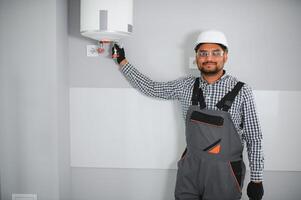 Indian plumber using screwdriver while fixing boiler or water heater, working on heating system in apartment photo
