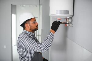 Indian plumber using screwdriver while fixing boiler or water heater, working on heating system in apartment photo