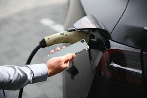 A man charges an electric car and checks the charge level on a smartphone photo