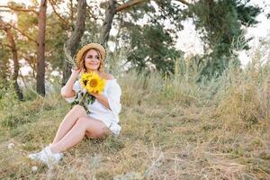 Outdoor shot of pleasant looking female with tanned healthy skin, dressed in white dress and summer hat, poses in park with confident satisfied expression, likes recreation. Beautiful young woman photo