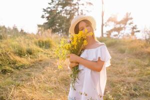 Outdoor shot of pleasant looking young girl with tanned healthy skin, dressed in white dress and summer hat, poses in park with confident satisfied expression, likes recreation. Beautiful young woman. photo