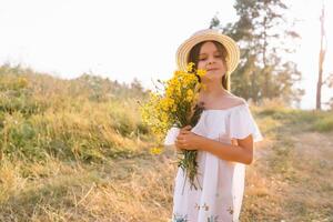 Outdoor shot of pleasant looking young girl with tanned healthy skin, dressed in white dress and summer hat, poses in park with confident satisfied expression, likes recreation. Beautiful young woman. photo