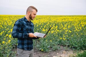 Young agronomist holds laptop in rape field. Agribusiness concept. agricultural engineer standing in a rape field with a laptop in summer. photo