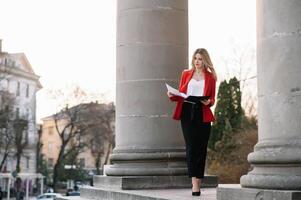 retrato de negocio mujer en sensación de concentrado estrés y ver estar y sostener el papel archivo sábana en el al aire libre peatonal caminar camino con el ciudad espacio de exterior moderno fachada edificio. foto