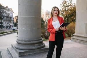 retrato de negocio mujer en sensación de concentrado estrés y ver estar y sostener el papel archivo sábana en el al aire libre peatonal caminar camino con el ciudad espacio de exterior moderno fachada edificio. foto