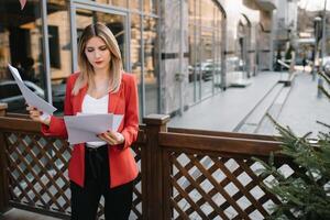 retrato de negocio mujer en sensación de concentrado estrés y ver estar y sostener el papel archivo sábana en el al aire libre peatonal caminar camino con el ciudad espacio de exterior moderno fachada edificio foto