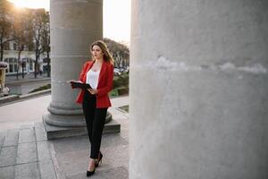 retrato de negocio mujer en sensación de concentrado estrés y ver estar y sostener el papel archivo sábana en el al aire libre peatonal caminar camino con el ciudad espacio de exterior moderno fachada edificio foto