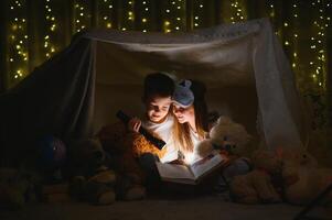 Little kids involving in reading amazing book. They lying in nice toy tent in playroom. Boy holding flashlight in hand photo