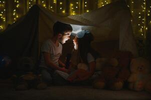 Two little child play at home in the evening to build a camping tent to read books with a flashlight and sleep inside. Concept of game, magic, creativity, alarm systems photo