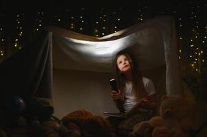 child girl reading with book and flashlight and teddy bear in tent. before going to bed photo