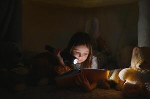 child girl reading with book and flashlight and teddy bear in tent. before going to bed photo