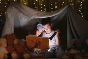 Two children with flashlight read a book under a blanket as a tent photo
