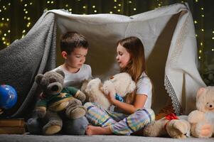 Two little children play at home in the evening to build a camping tent to read books with a flashlight and sleep inside. Concept of game, magic, creativity, alarm systems photo