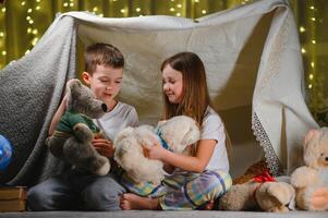 Two little child play at home in the evening to build a camping tent to read books with a flashlight and sleep inside. Concept of game, magic, creativity, alarm systems photo