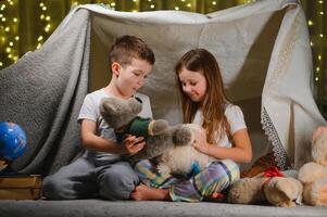Two little child play at home in the evening to build a camping tent to read books with a flashlight and sleep inside. Concept of game, magic, creativity, alarm systems photo