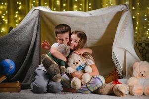 Two little child play at home in the evening to build a camping tent to read books with a flashlight and sleep inside. Concept of game, magic, creativity, alarm systems photo