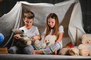 Two little children play at home in the evening to build a camping tent to read books with a flashlight and sleep inside. Concept of game, magic, creativity, alarm systems photo