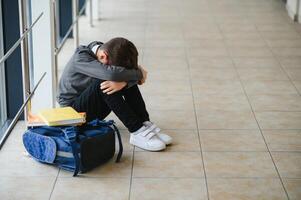 Little boy sitting alone on floor after suffering an act of bullying while children run in the background. Sad young schoolboy sitting on corridor with hands on knees and head between his legs photo