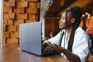 Beautiful african business woman waiting for the meeting in a co-working space and sitting with the portable computer. Hipster girl is surfing the web on a laptop during lunch. photo