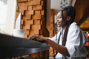 Beautiful african business woman waiting for the meeting in a co-working space and sitting with the portable computer. Hipster girl is surfing the web on a laptop during lunch. photo
