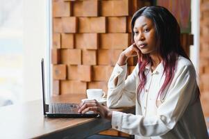 Beautiful african business woman waiting for the meeting in a co-working space and sitting with the portable computer. Hipster girl is surfing the web on a laptop during lunch. photo