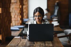 Beautiful african business woman waiting for the meeting in a co-working space and sitting with the portable computer. Hipster girl is surfing the web on a laptop during lunch. photo