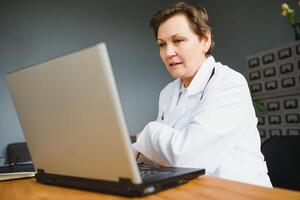 Portrait of female doctor counseling patient via call. Professional physician in white lab coat gesturing and explaining course of treatment sitting at office desk during online consultation. photo