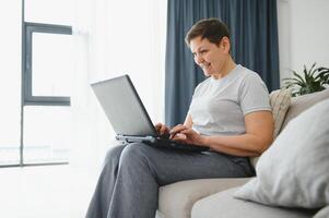 Elderly female sit on couch holding on lap laptop search information, check or typing e-mail, spend time on-line, modern tech, easy interesting pastimes services for retired older generation concept. photo