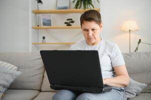 Elderly female sit on couch holding on lap laptop search information, check or typing e-mail, spend time on-line, modern tech, easy interesting pastimes services for retired older generation concept. photo