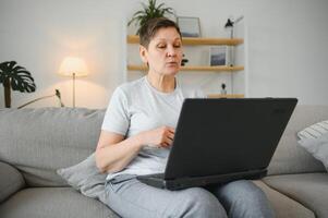 Older mature woman using wireless laptop apps browsing internet sit on sofa, smiling middle aged grandmother working distantly on computer surfing web communicating online looking at screen at home. photo