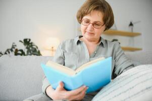 Restful reading. Portrait of thoughtful aged woman reading favorite literature at cozy home. She is lying on pillows on comfortable sofa. Legs are covered with soft blanket. photo