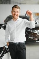 Happy buyer holding keys near the car in front of the modern avtosalon building photo