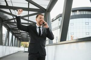 Businessman smiling and raising his fist in the air, with office building background - business success, achievement, and win concepts photo