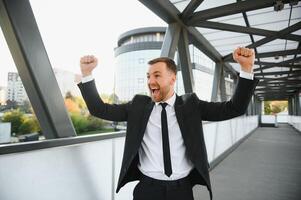 Businessman smiling and raising his fist in the air, with office building background - business success, achievement, and win concepts photo