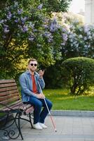 Visually impaired man with walking stick, sitting on bench in city park. Copy space photo