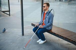 Visually impaired man with walking stick, sitting on bench in city park. Copy space photo