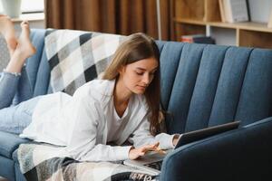 Smiling attractive young woman sitting on sofa using laptop communicating working online at home, happy teen girl typing on computer, enjoying writing blog or chatting with friends in social network photo