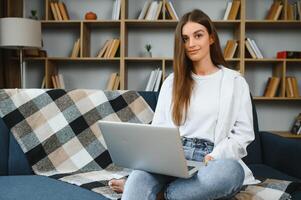 Smiling attractive young woman sitting on sofa using laptop communicating working online at home, happy teen girl typing on computer, enjoying writing blog or chatting with friends in social network photo