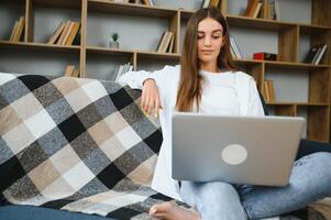 Smiling attractive young woman sitting on sofa using laptop communicating working online at home, happy teen girl typing on computer, enjoying writing blog or chatting with friends in social network photo