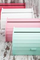 Colorful wooden bread boxes placed on a rustic white wooden surface. Bread boxes featuring a roll top opening mechanism with horizontal slats. photo
